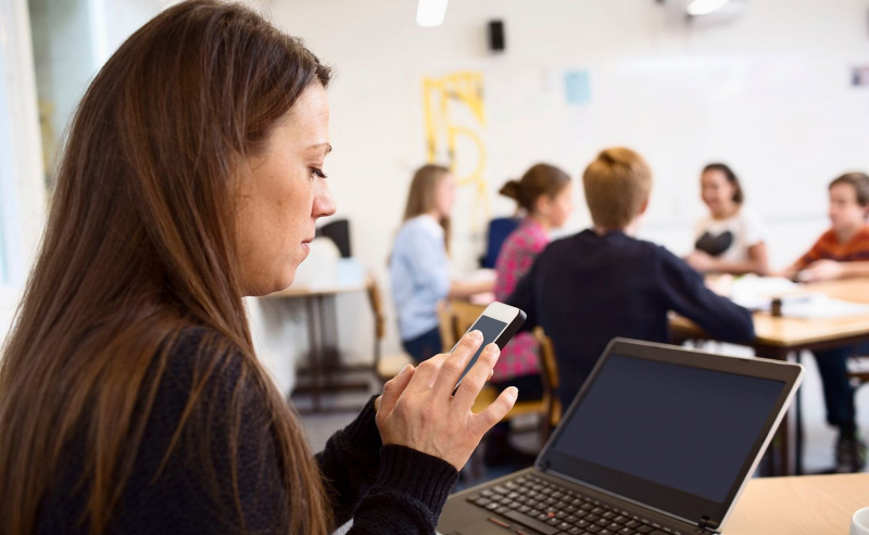 Profesor a través de teléfono móvil en el aula con estudiantes en segundo plano.
