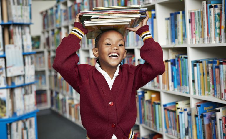 Scolara carina sorridente e in equilibrio pila di libri sulla testa in biblioteca