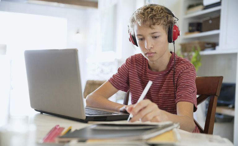 Jeune garçon prenant des notes sur un bloc-notes tout en utilisant un ordinateur portable et des écouteurs à table