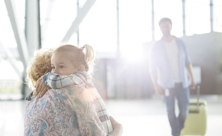 Madre e hija abrazándose en el aeropuerto con padre en segundo plano.
