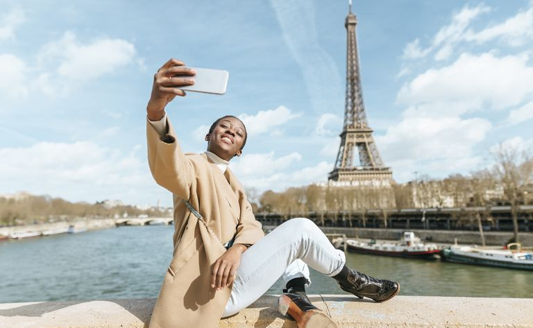 Francja, Paryż, Kobieta siedząca na moście nad Sekwaną na tle wieży Eiffla robi selfie