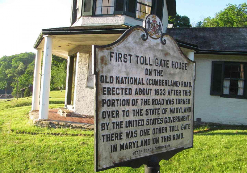 Casa de pedágio e marcador histórico na Estrada Nacional em um dia ensolarado.