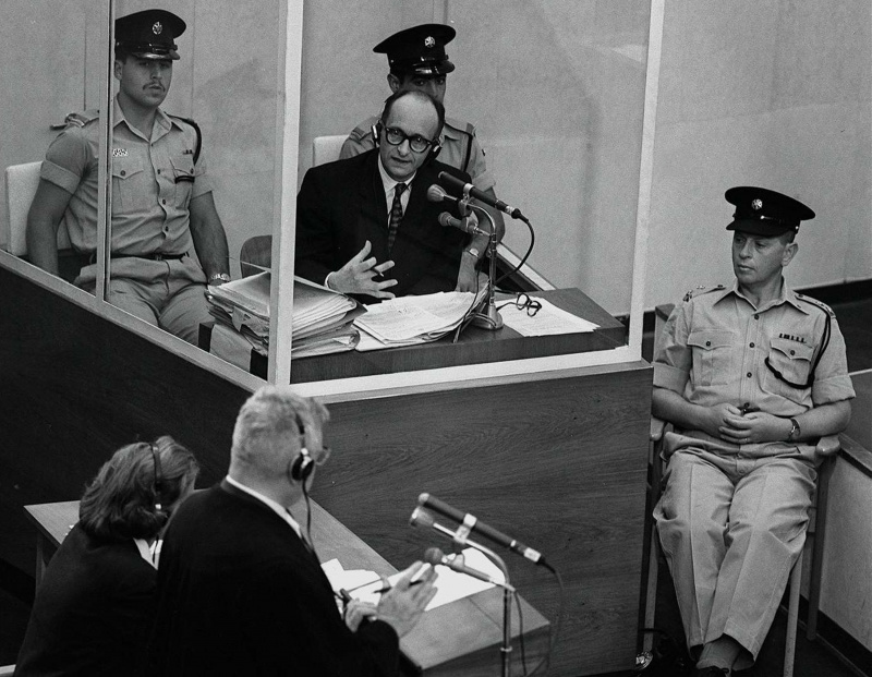 Nacistu kara noziedznieks Ādolfs Eihmans viņa tiesas prāvas laikā 1961. gada 22. jūnijā Jeruzalemē stāv stikla aizsargājošā kabīnē, kurai blakus atrodas Izraēlas policija.