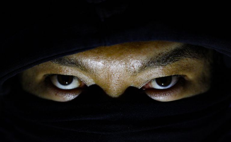 Um ninja nas sombras olhando por trás de uma máscara preta.