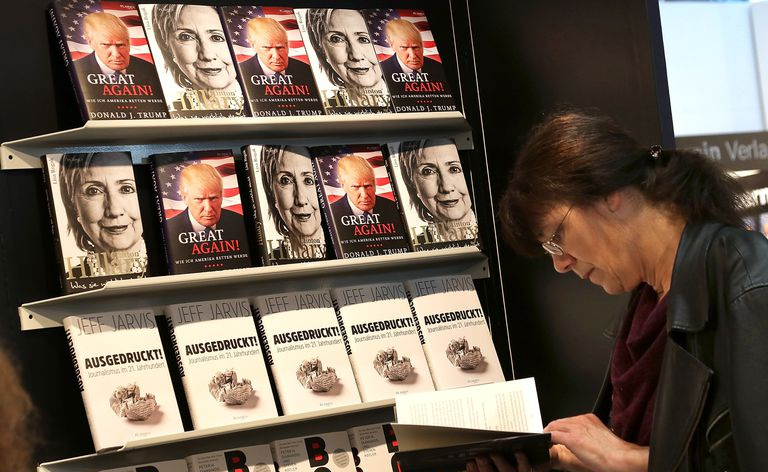 Una mujer mira un libro frente a una pantalla que muestra libros de Donald Trump y Hillary Clinton.