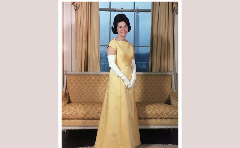 Lady Bird Johnson dzeltenā kleitā un garos baltos cimdos, pie loga - skats uz Vašingtonas pieminekli