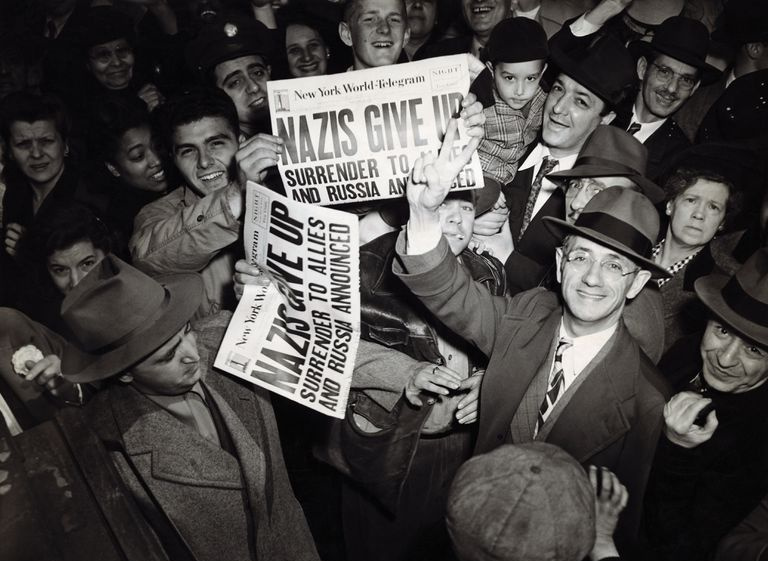 New Yorker feiern den V-E Day und das Ende des Zweiten Weltkriegs, halten Zeitungen und lächelnde Schwarzweißfotos hoch.