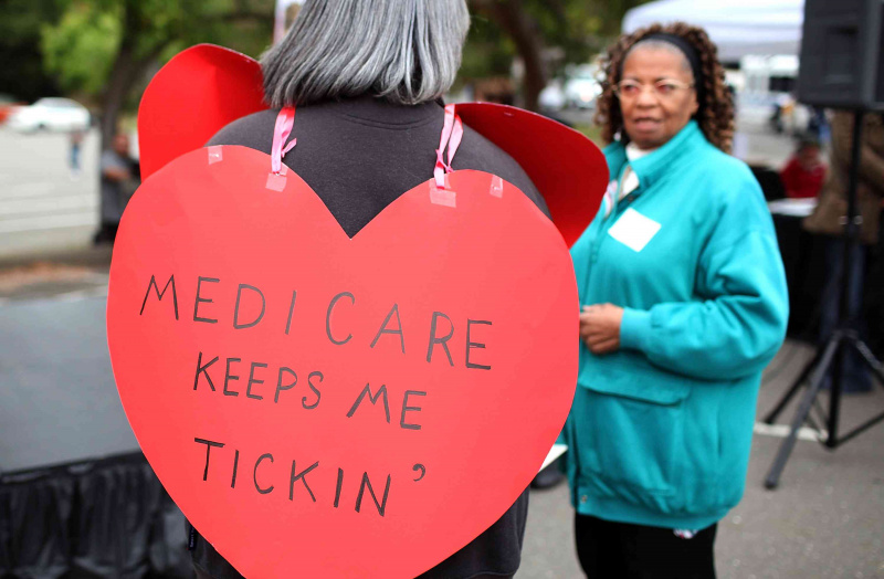 Sieviete ar sirds formas izkārtni ar uzrakstu “Medicare Keeps Me Ticking”