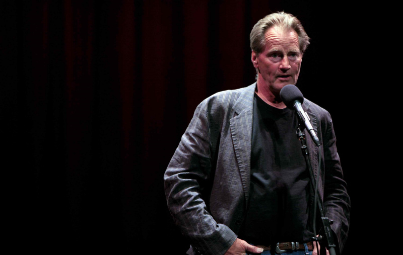 Sam Shepard, stojący z rękami w kieszeniach, przy mikrofonie