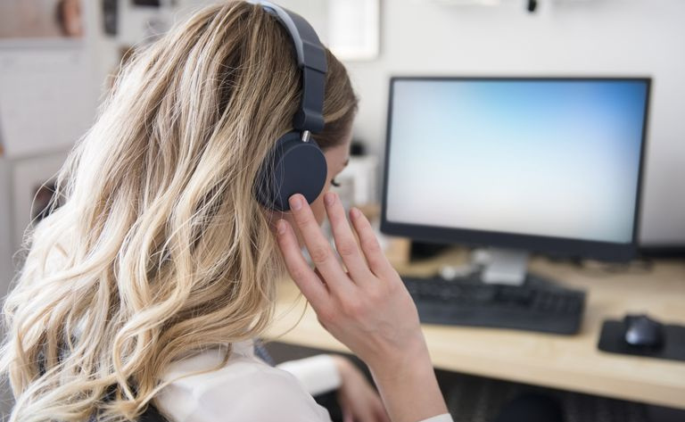 Kaukasisk kvinne lytter til hodetelefoner ved datamaskinens skrivebord