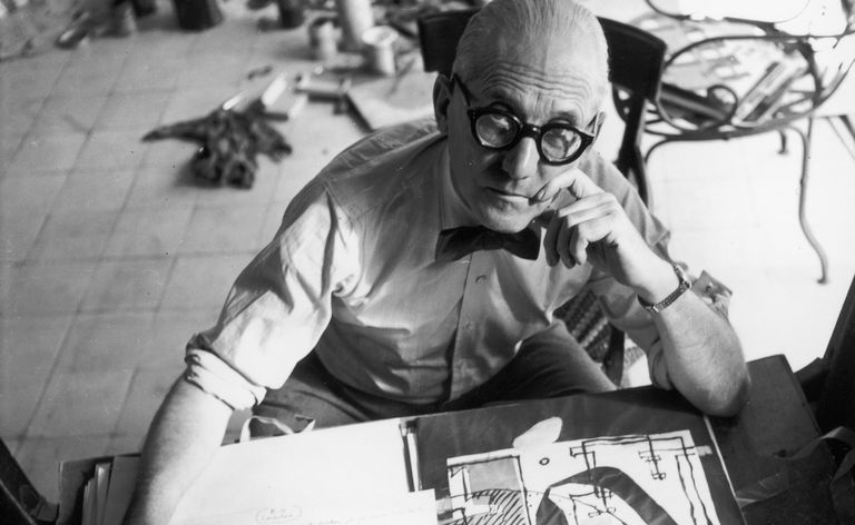 Den sveitsiskfødte arkitekten Le Corbusier, iført briller, sløyfe og skjorteermer, ser opp fra et tegnebord og en åpen portefølje